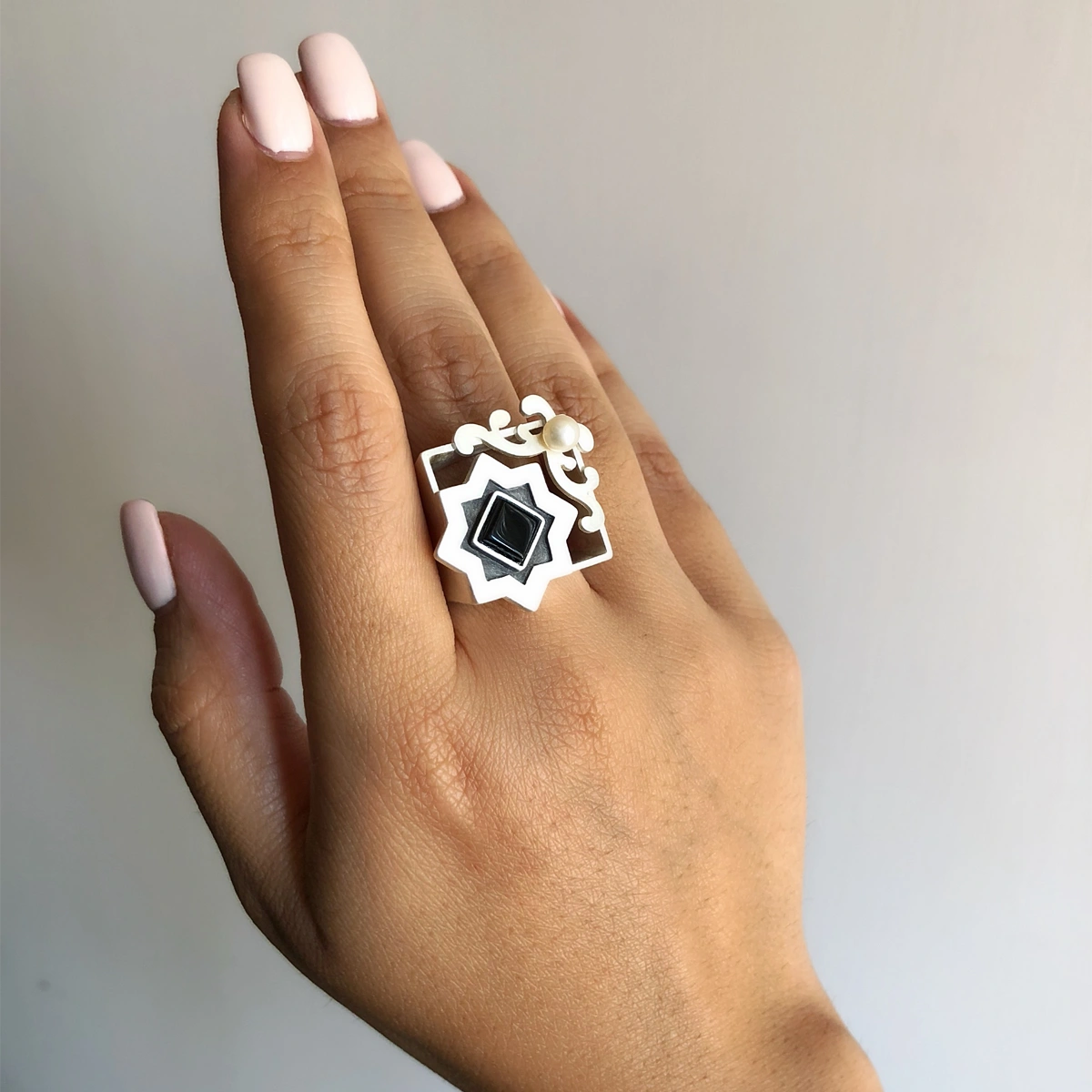 hand wearing handmade silver statement ring with geometric star design, square black onyx centerpiece, freshwater pearl accent, and oxidized details.