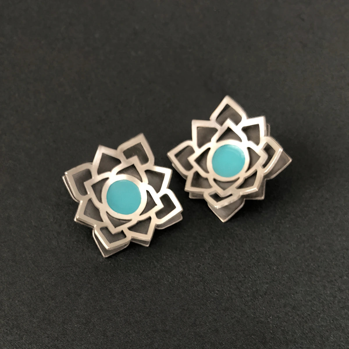Handmade sterling silver lotus stud earrings with layered openwork design, oxidized silver base, and turquoise-blue enamel centerpiece.