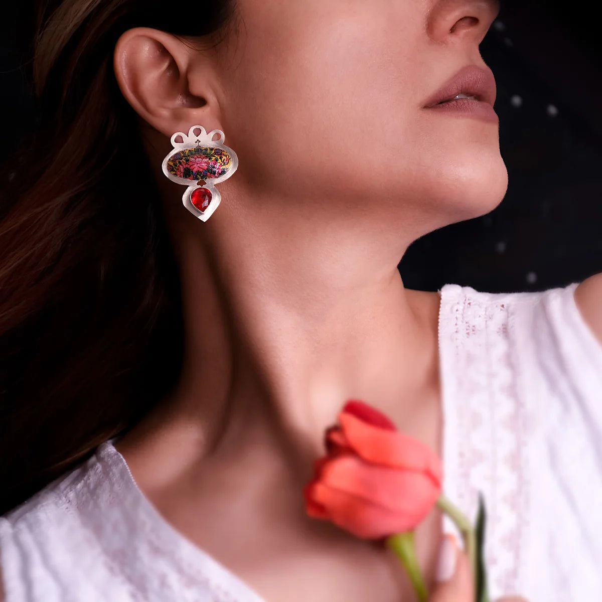 Woman wearing handmade silver statement stud earrings in oval shape, decorated with hand-printed floral patterns and tear-drop red cubic zirconia crystals. 