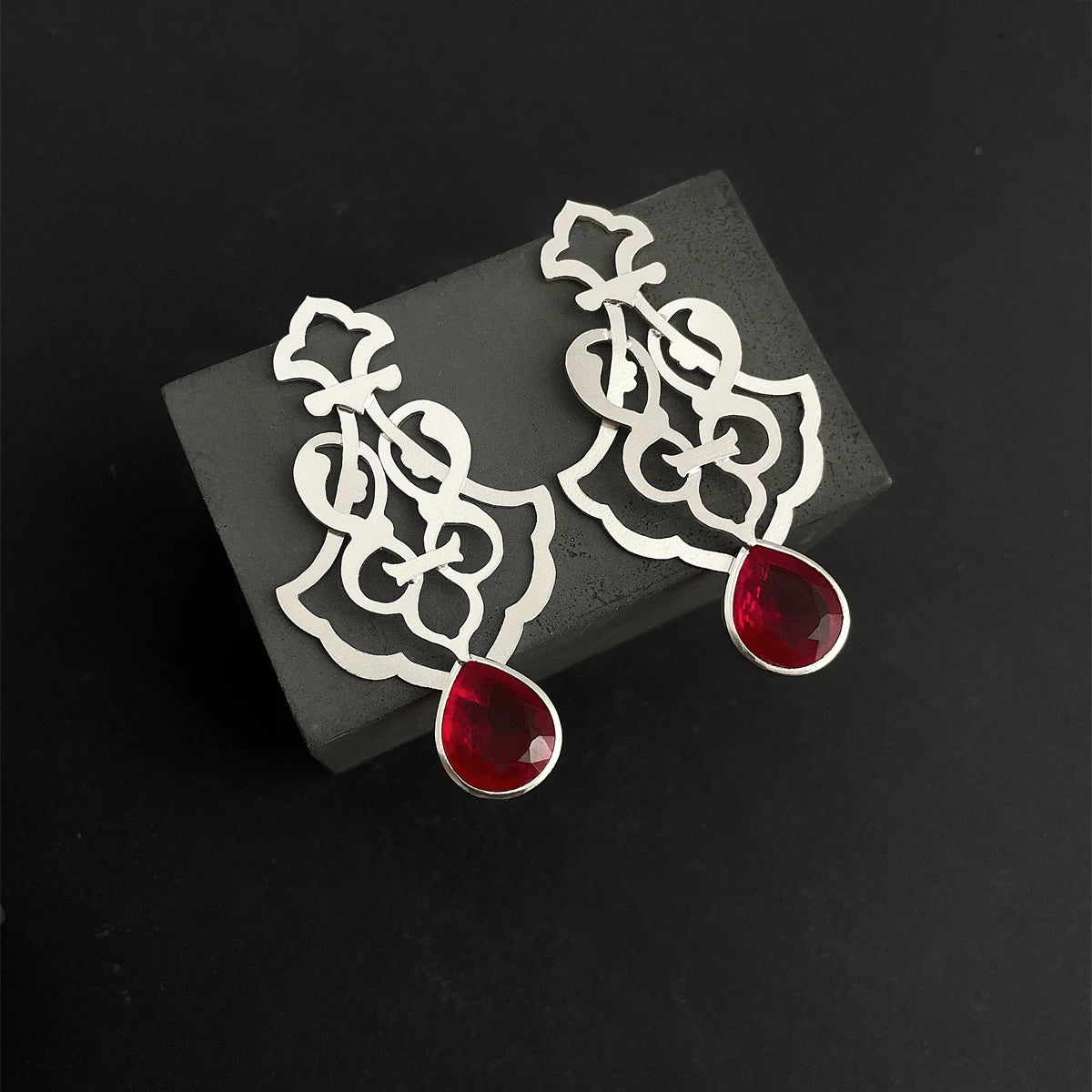 Handmade sterling silver openwork earrings with interwoven floral patterns and faceted teardrop red crystals.