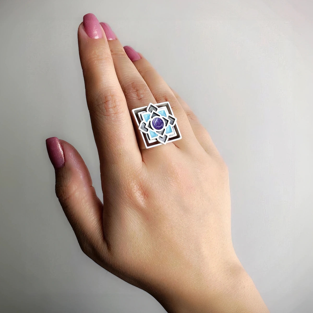 Hand wearing Handmade sterling silver statement ring with square band, geometric lotus design,  purple and blue enamel detailing.